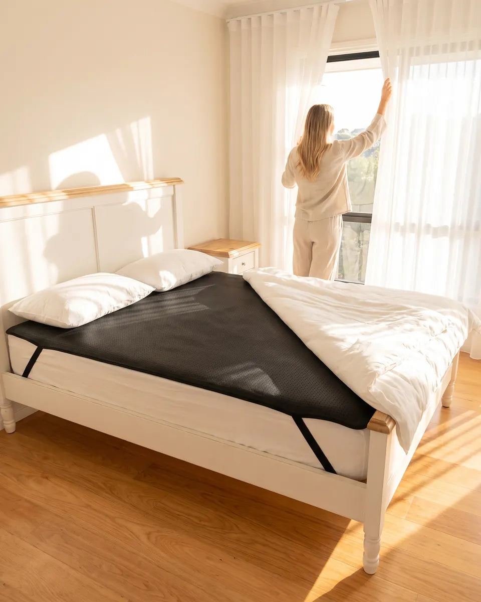 A woman opens the curtains to morning light; the GroundingFlow mat covers the bed